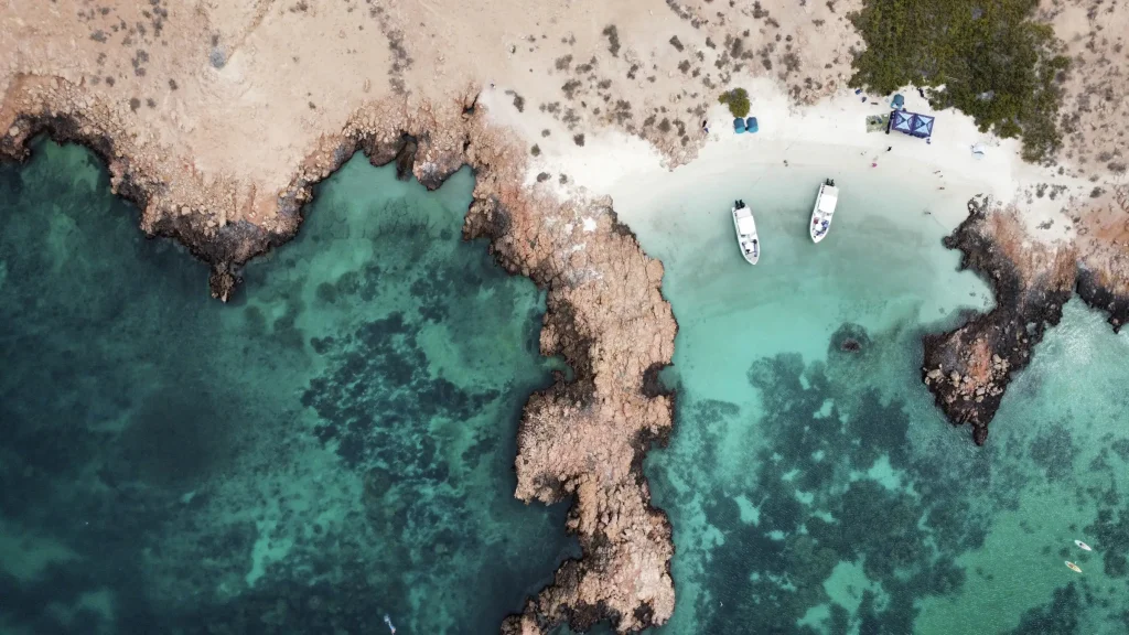 Aerial view of two boats anchored beside a white sand beach with turquoise water and coral reef at the Daymaniyat Islands, Oman