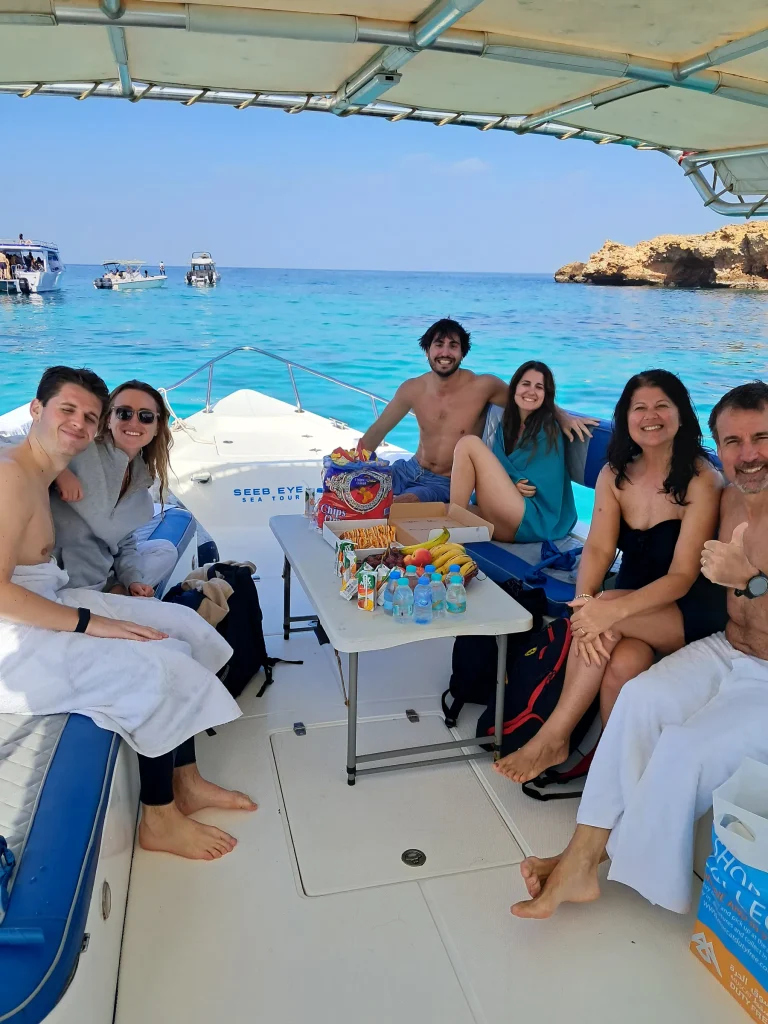 Group of guests sitting around a table of fresh fruit on the shaded boat deck, with turquoise water and an island behind